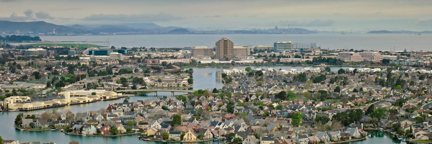 Aerial view of a cityscape with residential neighborhoods and water bodies, overlooking a bay and distant mountains.